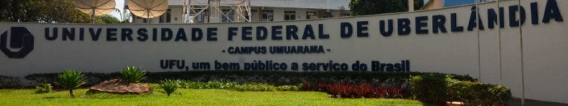 A imagem mostra a entrada do campus Umuarama da Universidade Federal de Uberlândia (UFU), com um grande letreiro e um jardim bem cuidado. Aqui estão alguns detalhes:  Letreiro: Exibe “UNIVERSIDADE FEDERAL DE UBERLÂNDIA - CAMPUS UMUARAMA” em letras brancas sobre um fundo escuro. Frase: Abaixo do nome, lê-se “UFU, um bem público a serviço do Brasil”. Jardim: Grama verde e arbustos vermelhos e verdes, indicando manutenção cuidadosa. Fundo: Antenas parabólicas grandes e uma torre de transmissão são visíveis. O 