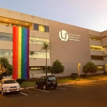 A imagem mostra um edifício moderno de vários andares, com uma fachada de cores claras. Na lateral do prédio, há uma grande faixa de tecido com as cores do arco-íris, simbolizando a diversidade e os direitos LGBTQ+. À frente do prédio, há uma área de estacionamento com dois carros estacionados. O logo da Universidade é visível na parte superior direita da imagem, e a iluminação do local sugere que a foto foi tirada ao entardecer.